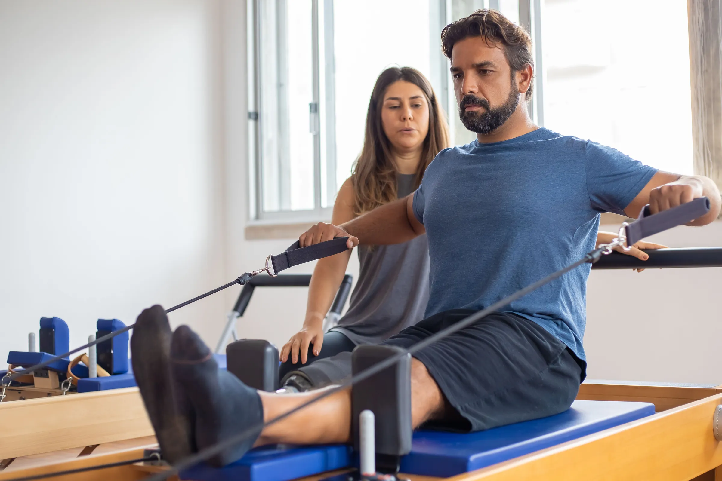A trainer guides a client through a Pilates reformer exercise in a bright studio